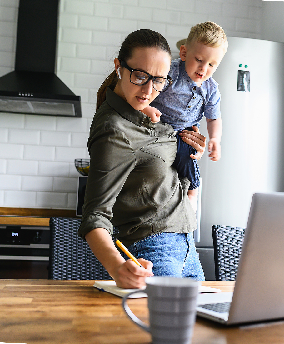 Woman standing at a kitchen counter with a child on her hip, writing in a notebook