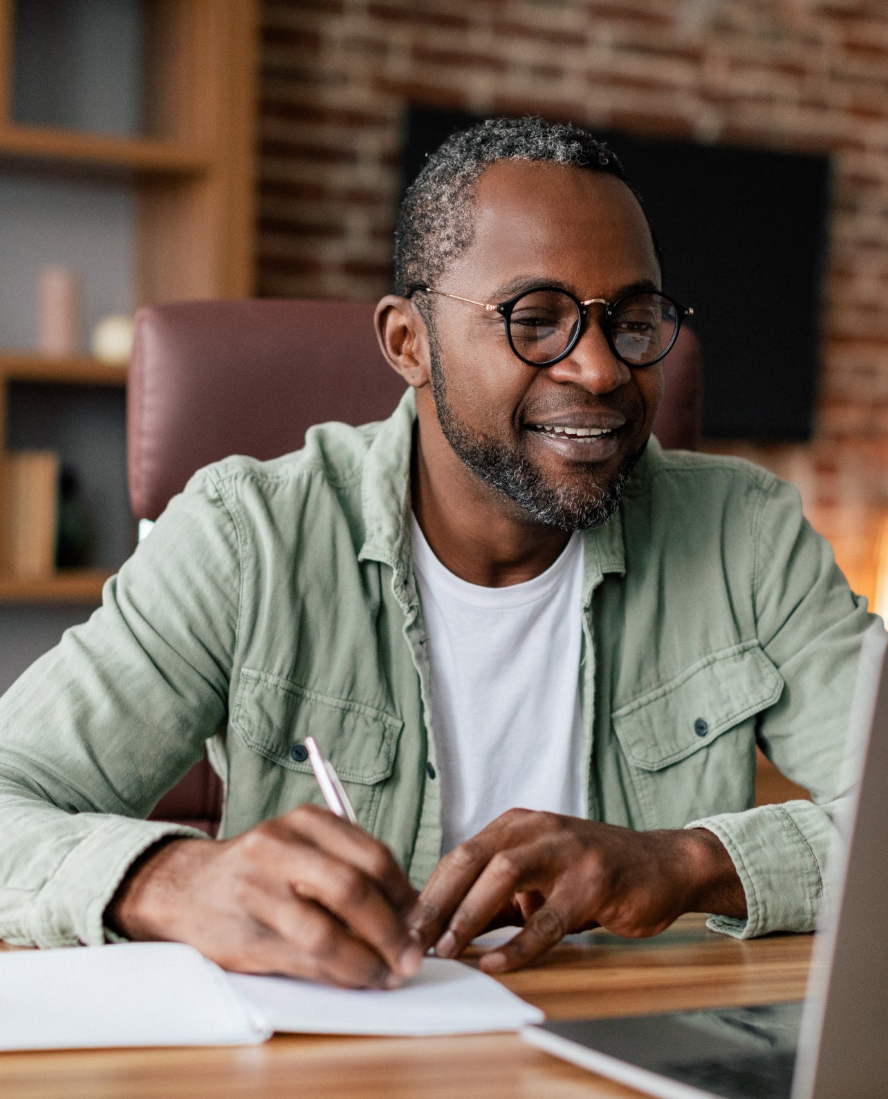 Man sitting at a desk with a laptop, writing in a notebook, in an office setting.