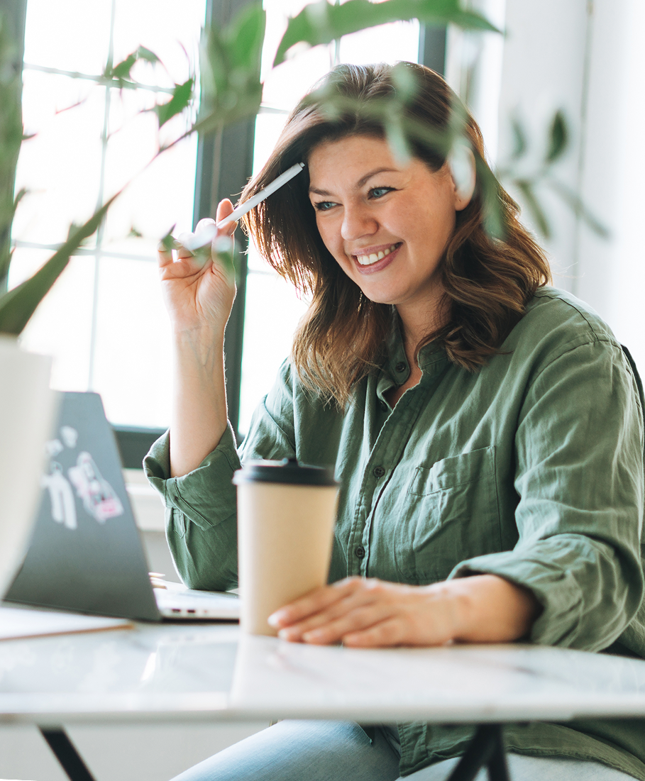 Woman sitting at a desk with a laptop, holding a coffee cup and a plant in the foreground.
