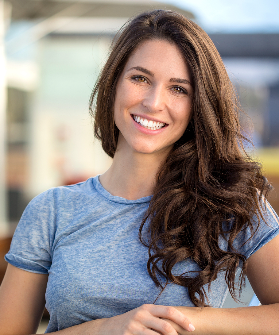 Woman with long brown hair wearing a blue shirt, smiling outdoors.