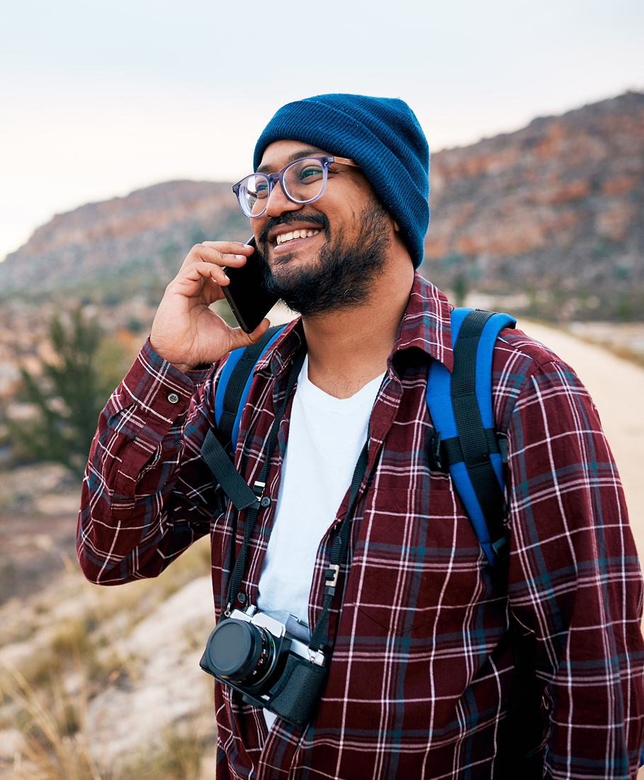 Man on a phone call with a backpack and camera, standing in a scenic outdoor setting.