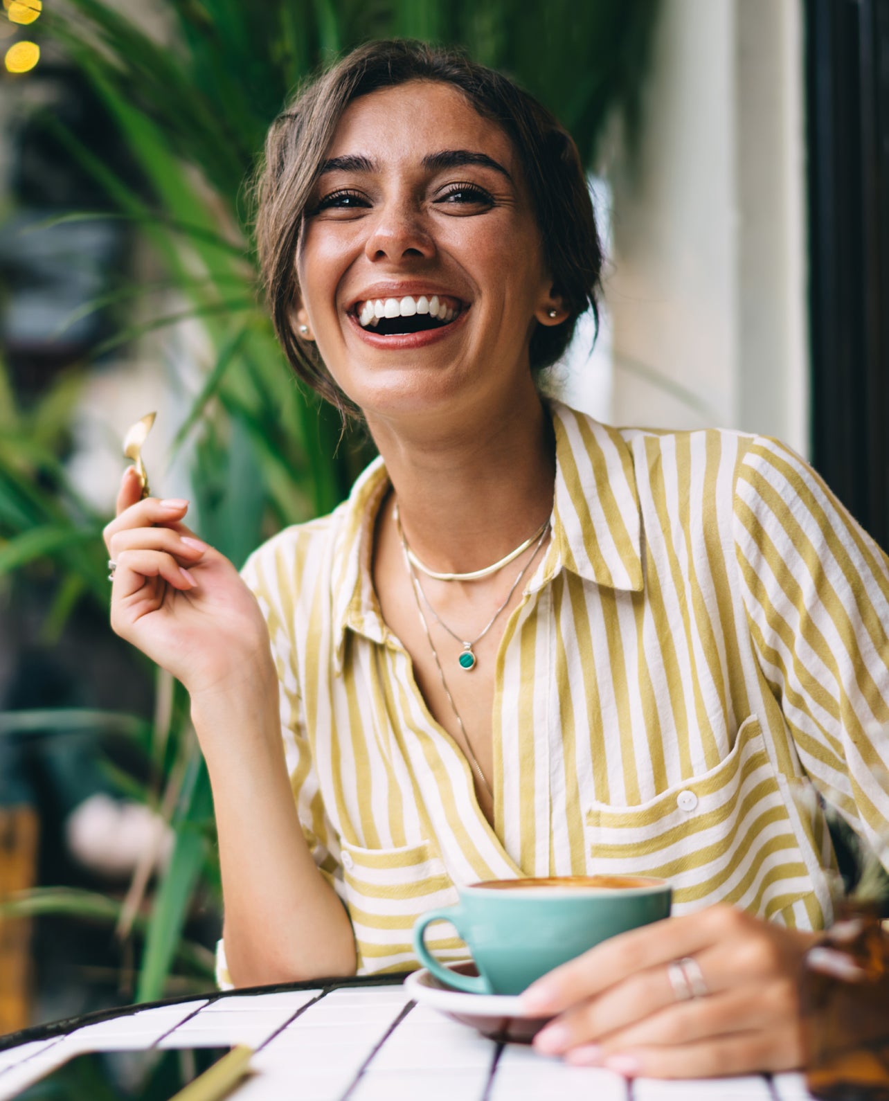 Woman in a striped shirt holding a cup and a spoon, smiling outdoors.