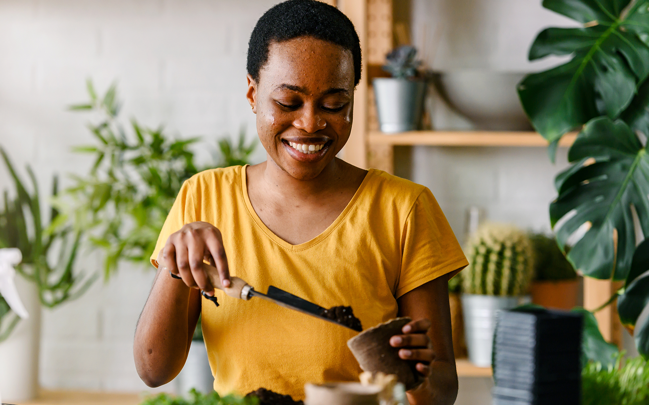 Woman in a yellow shirt holding a plant in a home setting with plants and decor.