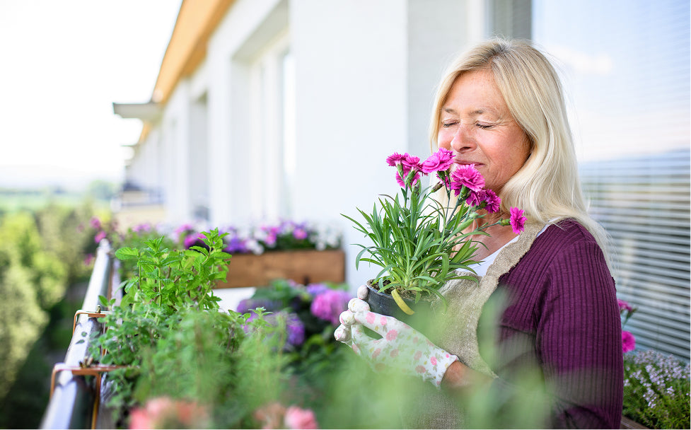Woman holding a potted plant with pink flowers on a balcony filled with gardening supplies.