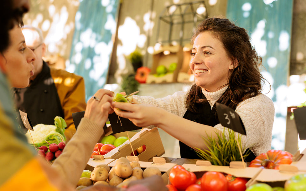 Woman at a farmers market interacting with another person over produce.