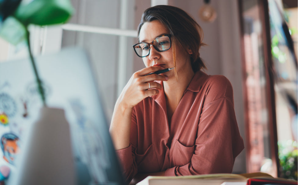 Woman wearing glasses and a pink shirt sitting at a desk with a laptop.
