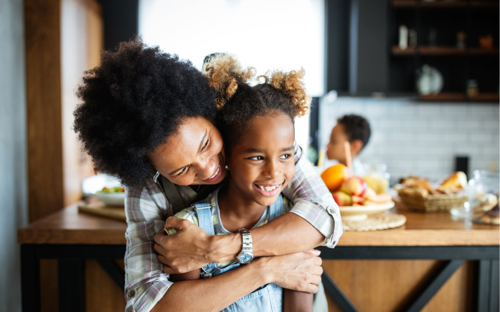 Woman hugging a young girl in a kitchen with fruit on the counter