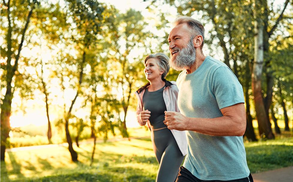 Man and woman jogging together in a park with trees and sunlight filtering through.