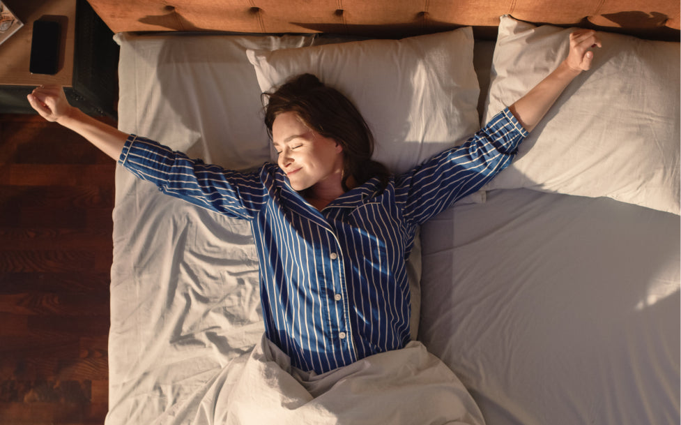 Woman waking up in bed with arms outstretched, wearing a blue striped pajama top.