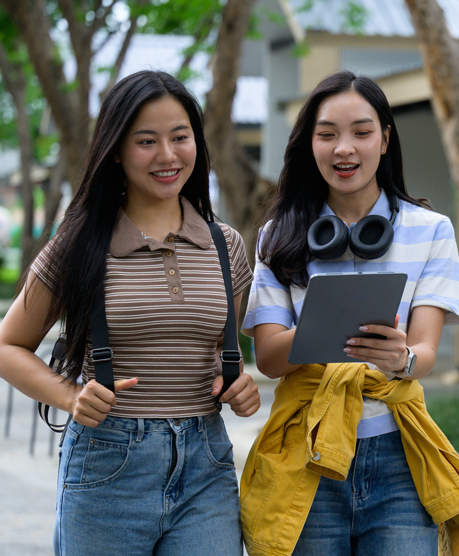 Two young women walking outdoors, one holding a tablet and headphones, smiling.