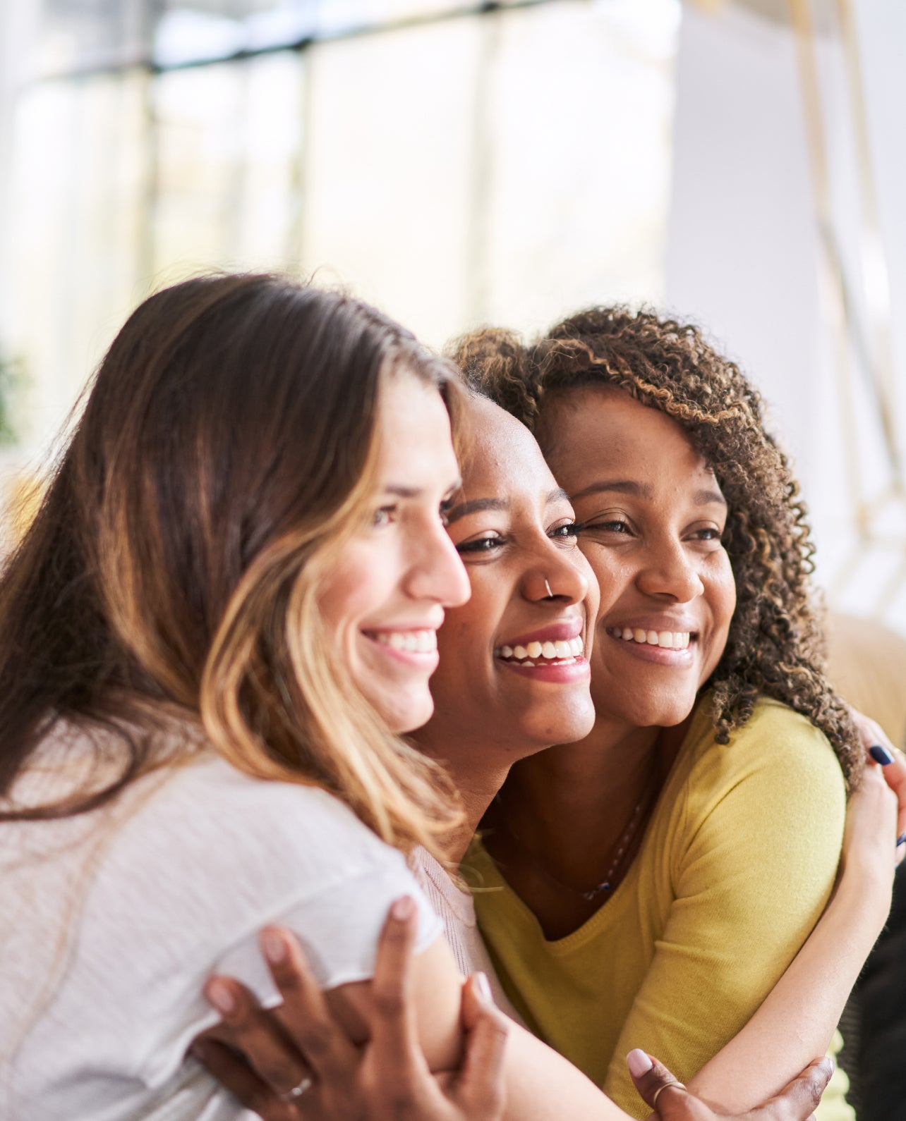 Three women hugging and smiling together as if posing for a photo in a bright indoor setting