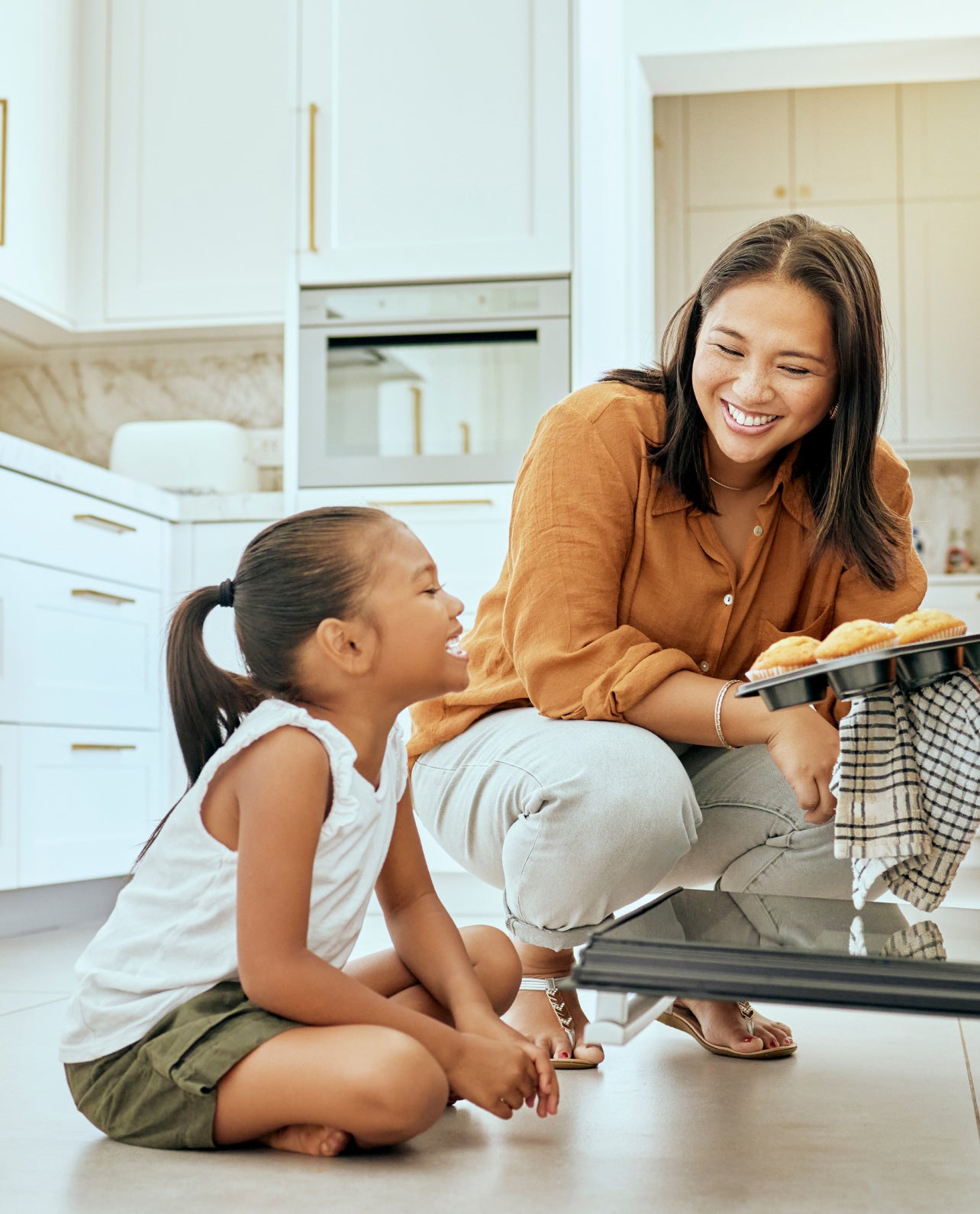 Woman and child in a kitchen taking a tray of muffins out of an oven.