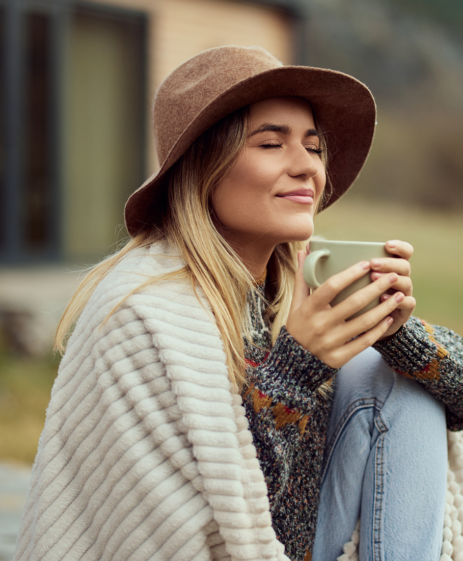 Woman wearing a brown hat and wrapped in a white blanket, holding a mug outdoors.