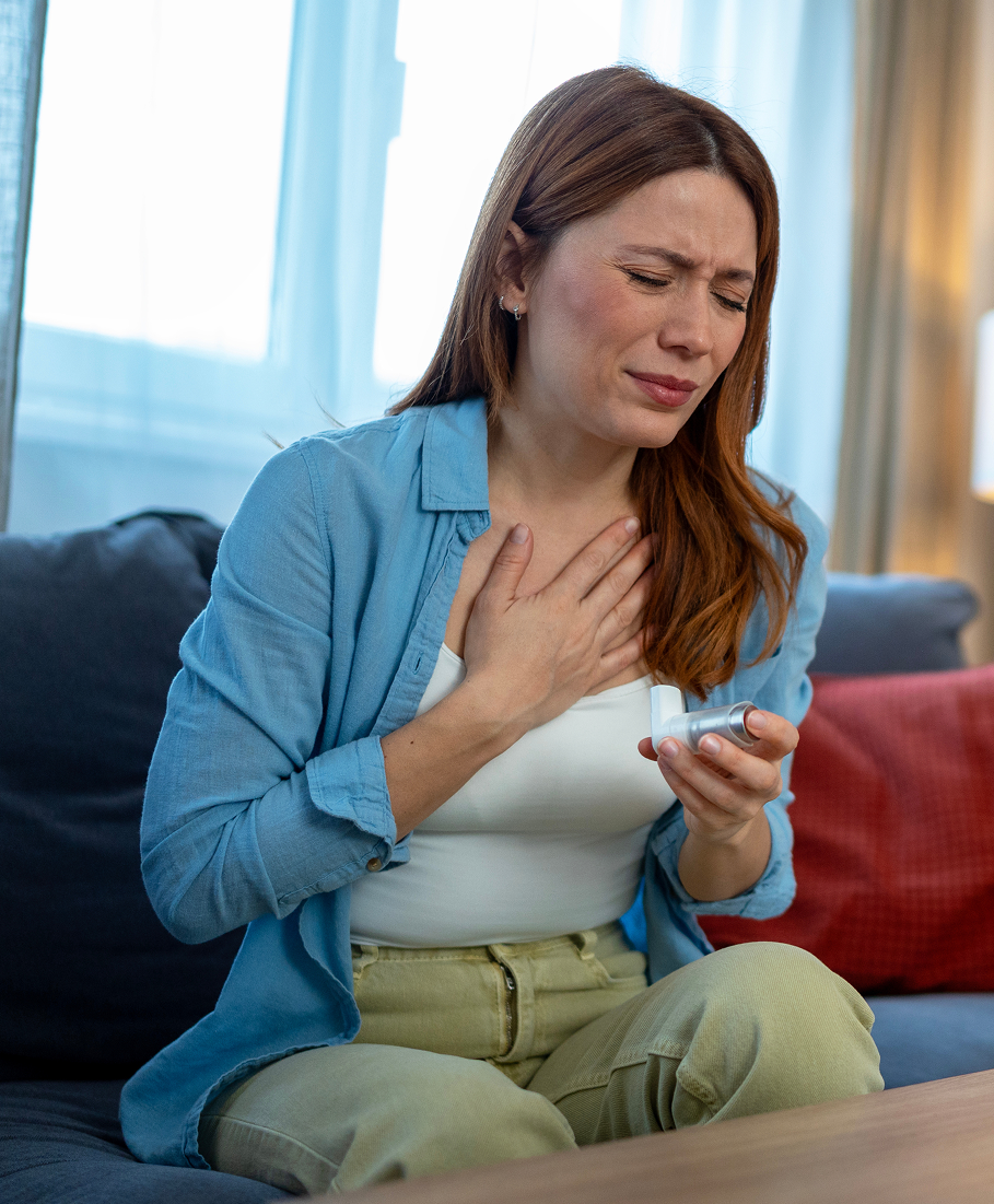 Woman sitting on a couch holding an inhaler and looking uncomfortable.