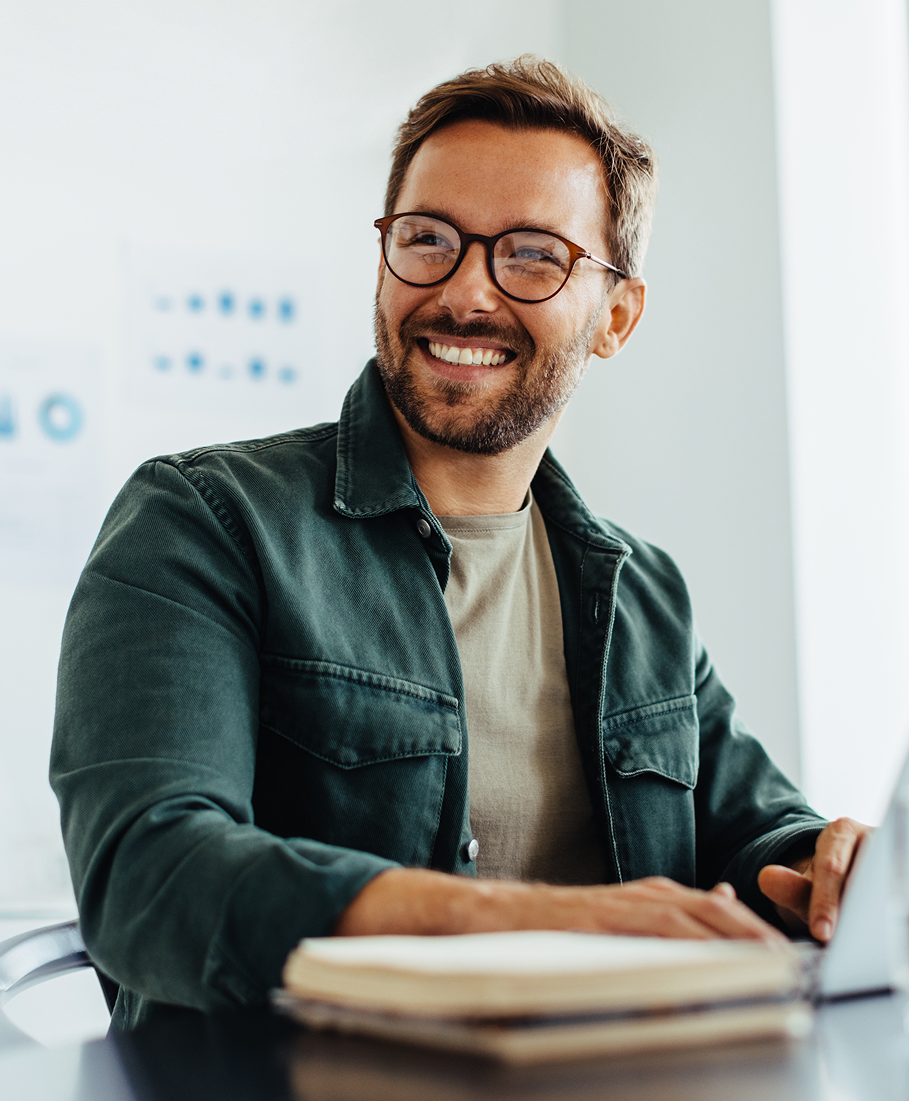 Man with glasses and a beard smiling at a desk with a notebook and laptop.