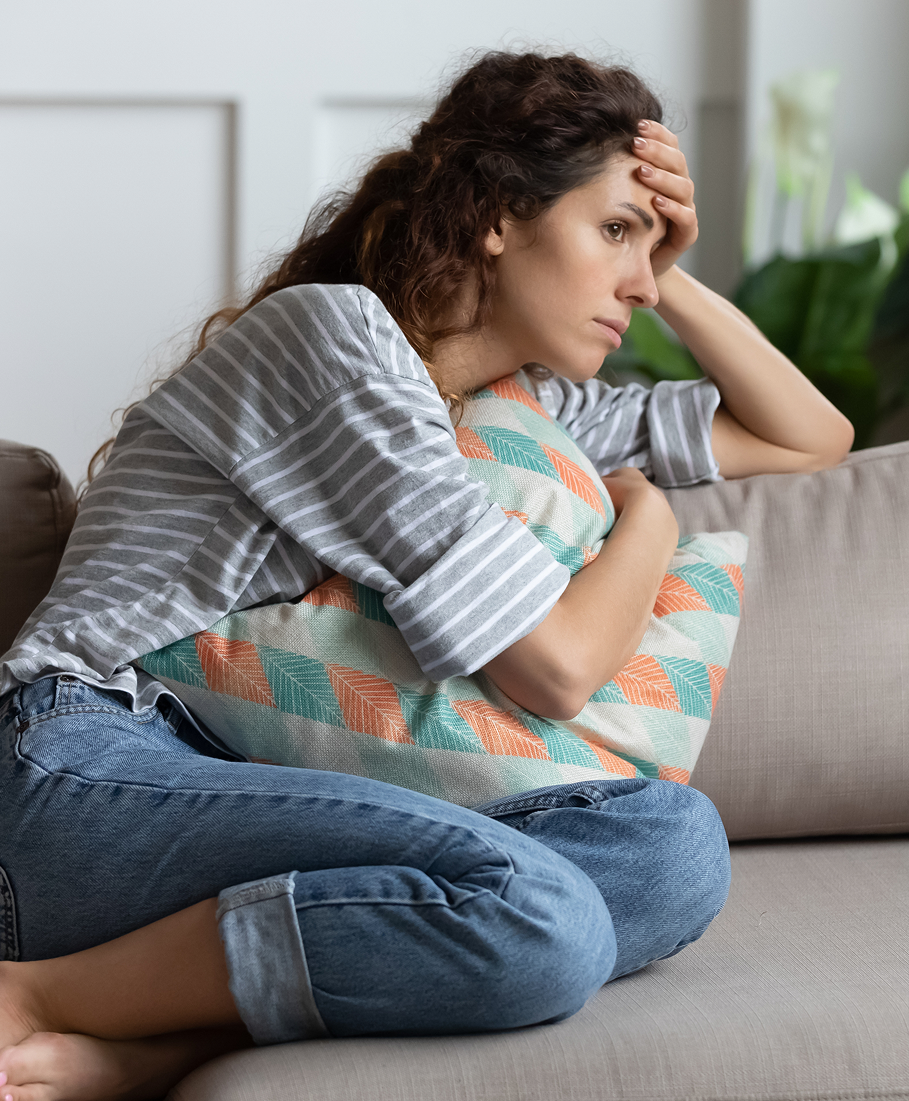 Woman sitting on a couch with a sad expression, holding her head and hugging a pillow to her stomach.
