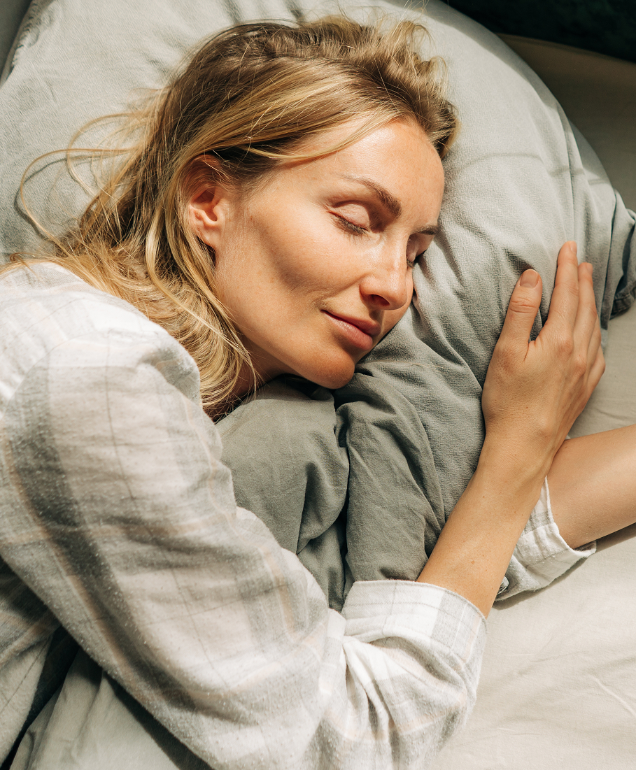 Woman lying in bed with gray bedding, eyes closed.