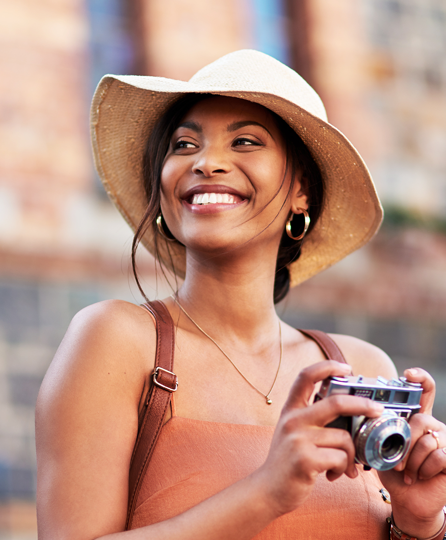 Woman wearing a wide-brimmed hat and holding a camera, smiling outdoors.