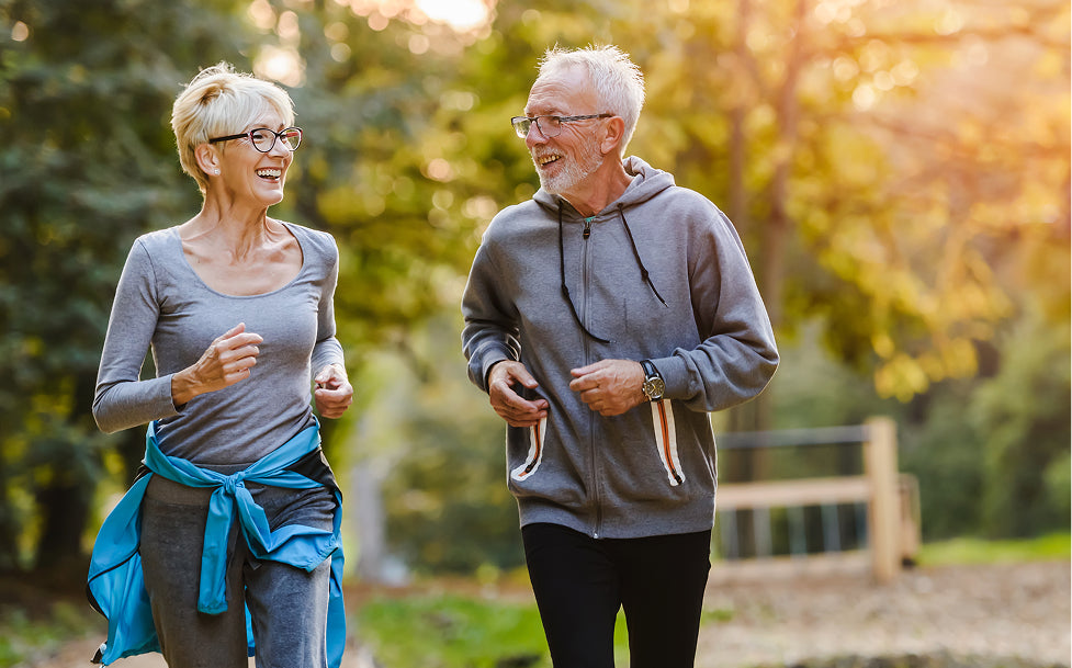 Two elderly people jogging together in a park with trees and sunlight filtering through.