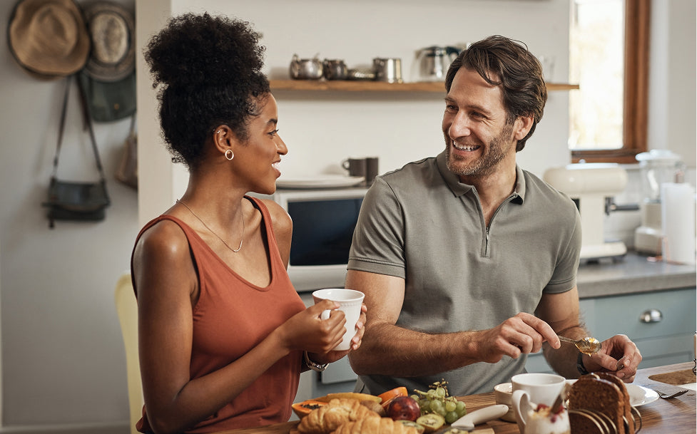 Man and woman sitting at a kitchen table with food and drinks, smiling and enjoying each other's company.