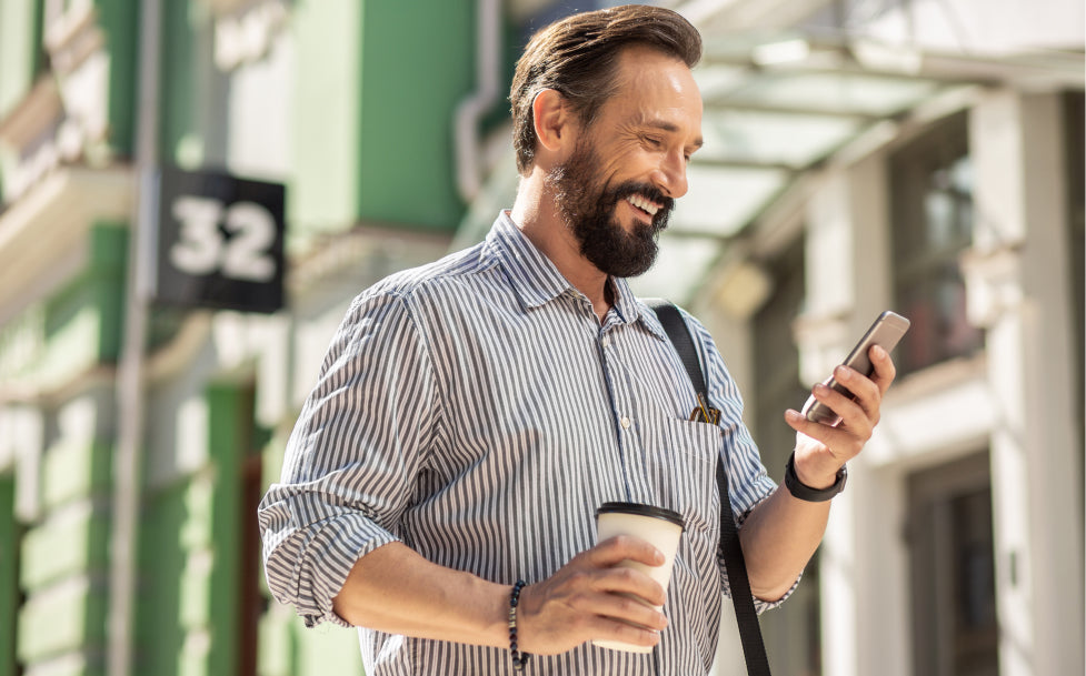 Man with a beard holding a coffee cup and smartphone, walking outdoors.