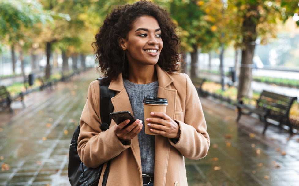 Woman walking outdoors holding a coffee cup and phone, surrounded by trees with autumn leaves.